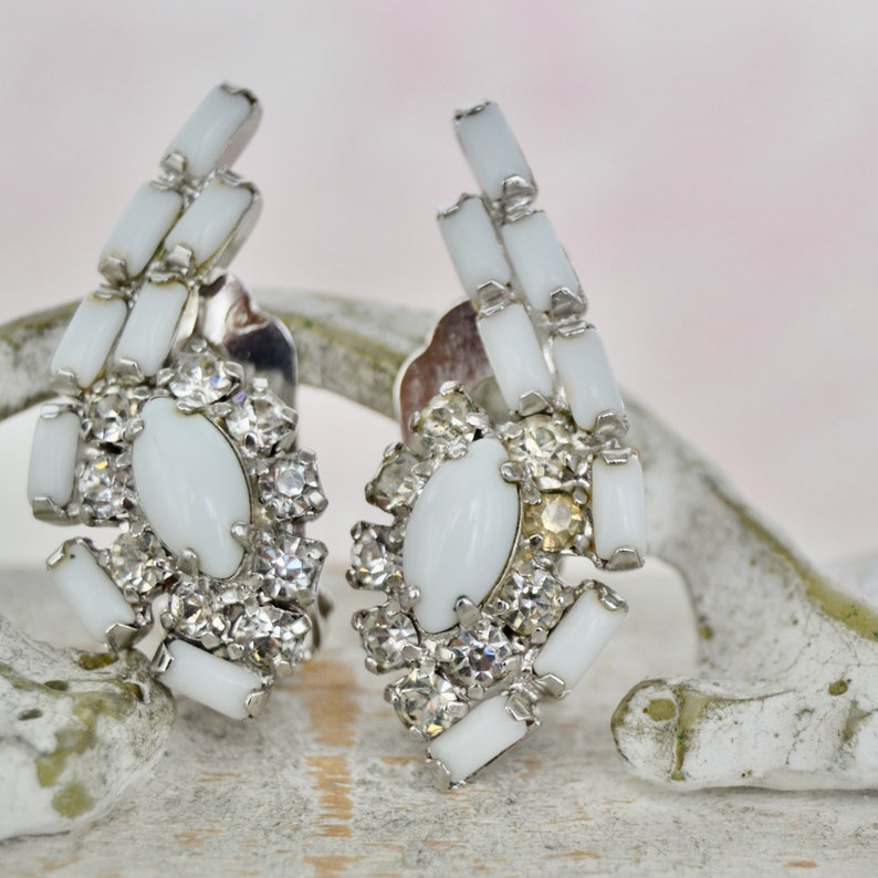 May include: A pair of white clip-on earrings with a cluster of clear rhinestones and a white oval stone in the center. The earrings are made of metal and have a vintage style.