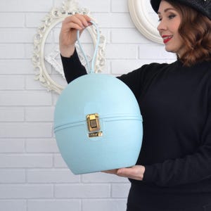 May include: A light blue, spherical vintage vanity case with a matching handle. The case has a textured surface and a gold-colored clasp. The handle is held by a person wearing a black sweater.