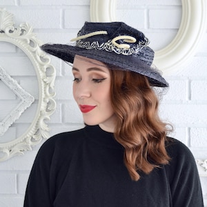 May include: A navy blue hat with a wide brim, adorned with a cream-colored rope and a delicate beaded trim. The hat features a sheer, dark veil and is set against a white brick wall.