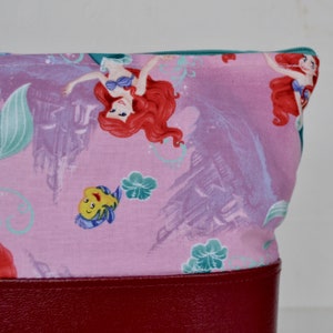 Puede incluir: Bolsa de tela rosa con cierre de cremallera y fondo de cuero rojo. La tela presenta un patr&oacute;n repetido de Ariel la Sirenita, Flounder y conchas marinas.