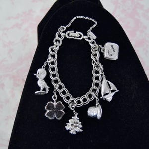 May include: Silver charm bracelet with various charms, including a duck, four-leaf clover, Christmas tree, thimble, sailboat, and a charm that says "FRANCE". The bracelet is displayed on a black surface.