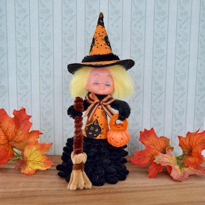 May include: A vintage Halloween witch doll with a black and orange outfit, holding a broom and a pumpkin-shaped treat bucket. The witch has blonde hair and a pointed hat with star accents. Decorative fall leaves are placed on either side.