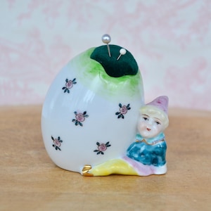 May include: A white porcelain pincushion shaped like an egg with a green top and pink floral accents. A small porcelain figurine of a jester is leaning against the pincushion.