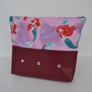 Puede incluir: Una bolsa rosa con cremallera y fondo burdeos. La bolsa presenta un patr&oacute;n repetido de Ariel, la Sirenita, nadando en el oc&eacute;ano.