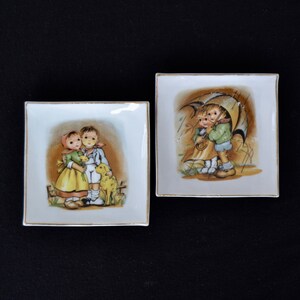 May include: Two square porcelain plates with gold trim. One plate features a painting of two children with a dog, while the other plate features a painting of two children under an umbrella.