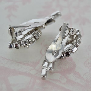May include: A pair of silver tone clip-on earrings with clear rhinestone accents. The earrings are in the shape of a teardrop with a cluster of rhinestones at the bottom.