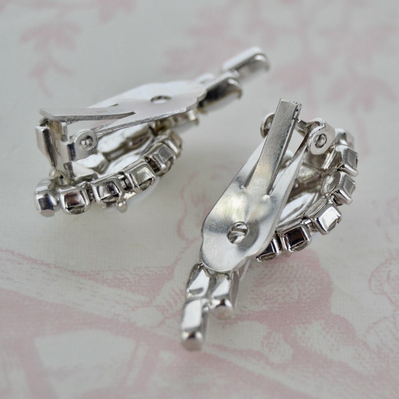 May include: A pair of silver tone clip-on earrings with clear rhinestone accents. The earrings are in the shape of a teardrop with a cluster of rhinestones at the bottom.