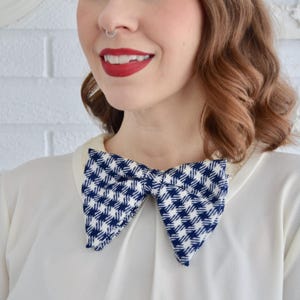 May include: A blue and white houndstooth bow tie. The bow tie is worn over a white collared shirt. The background is a white brick wall.