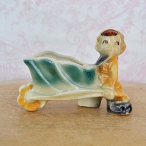 May include: A ceramic planter shaped like a boy pushing a wheelbarrow. The planter is painted in shades of green, yellow, and brown. The boy has a brown hat and a brown face.