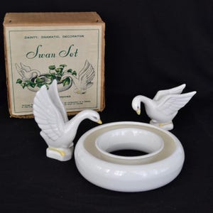 May include: A white ceramic swan salt and pepper shaker set. The swans are perched on a white ceramic ring. The set comes in a brown box with the words "Swan Set" printed on the front.