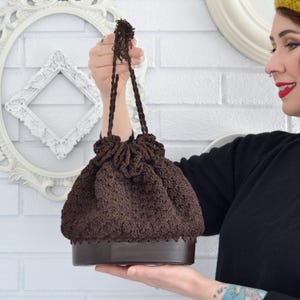 May include: A brown crocheted drawstring bag with a brown leather bottom. The bag has a gathered top and a long, brown crocheted handle.