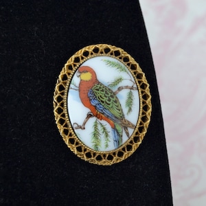 May include: Oval brooch with a gold-coloured frame, featuring a painted bird design. The bird has red, green, and blue feathers and sits on a branch. The brooch has an openwork border.
