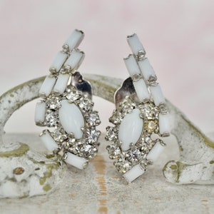 May include: A pair of white clip-on earrings with a cluster of clear rhinestones and a white oval stone in the center. The earrings are made of metal and have a vintage style.