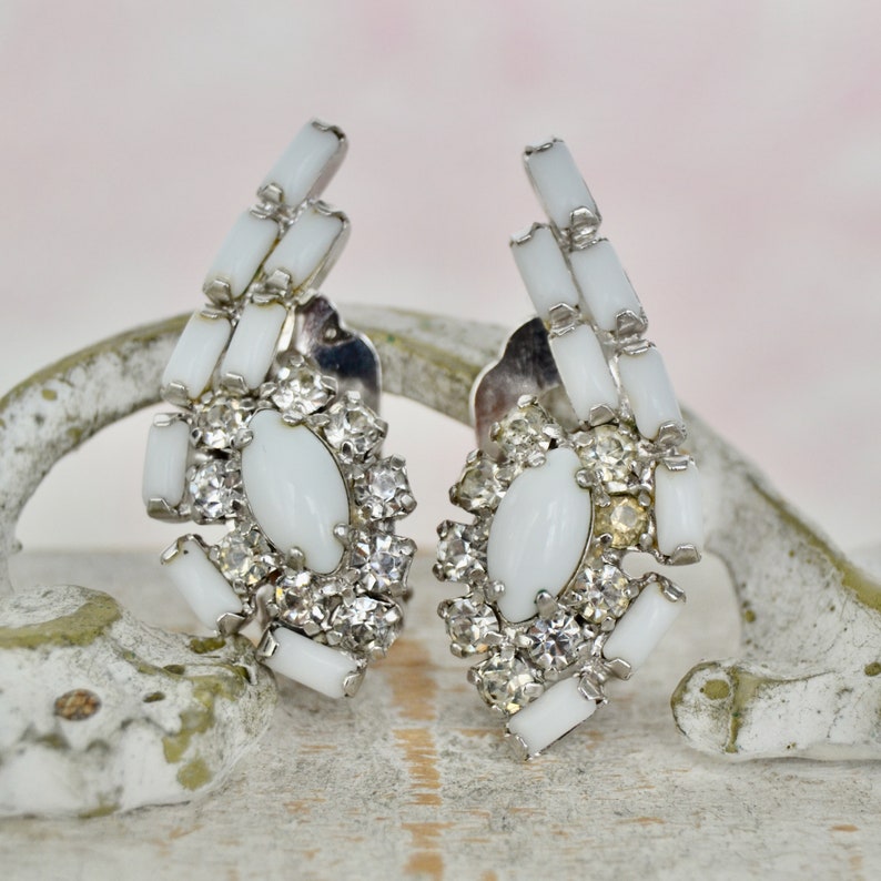 May include: A pair of white clip-on earrings with a cluster of clear rhinestones and a white oval stone in the center. The earrings are made of metal and have a vintage style.