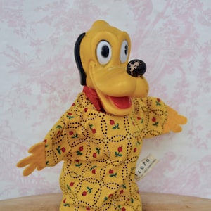 May include: A yellow plush puppet of Pluto the dog wearing a yellow and red dress with a floral pattern. The puppet has a black nose and large, black eyes.