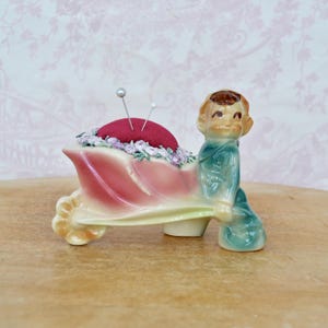 May include: A ceramic figurine of a child pushing a pink wheelbarrow that serves as a pincushion. The wheelbarrow contains a deep red velvet cushion surrounded by small purple flowers. Two silver sewing pins are inserted into the red cushion.