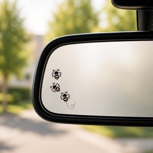 May include: Black and yellow bee stickers are arranged on a car mirror. The stickers are in a whimsical design, with three bees in a cluster and one bee with a dotted flight path. The mirror is framed in black.