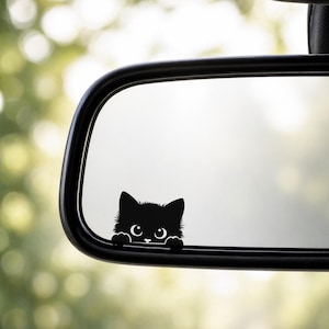May include: A black cat decal peeking over a car's rearview mirror. The cat has large, round eyes and a fluffy appearance. The mirror is black and has a curved shape, with a blurred green background.