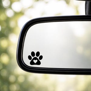 May include: Black paw print sticker with a heart-shaped design, placed on a car's rearview mirror. The sticker is a simple, yet charming, way to show love for pets. The background is blurred, with a green and white bokeh effect.