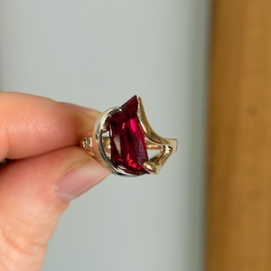 May include: A gold-toned ring featuring a large, rectangular, deep red gemstone. The gemstone is set in a unique, open-work design with a curved, asymmetrical shape. The ring's band has delicate detailing.