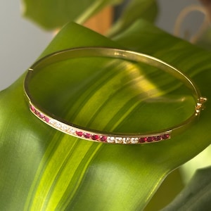 May include: A gold-colored bangle bracelet with a row of small, red gemstones and clear crystals. The bracelet is set against a green leaf, creating a contrast of colors. The bracelet appears to be made of metal.