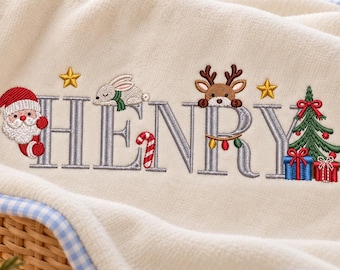 Op de afbeelding: Een crèmekleurige babydeken met een blauw-witte vichy-ruitrand. De deken is voorzien van geborduurde grijze letters die de naam HENRY vormen, versierd met een kerstman, een konijn, een rendier, een zuurstok, sterren, een kerstboom en cadeautjes.