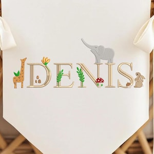 May include: A cream-colored banner with the name "DENIS" embroidered in gold, featuring animal and plant illustrations. A giraffe, elephant, mushroom, and rabbit are included in the design. The banner is tied with cream-colored ribbons.