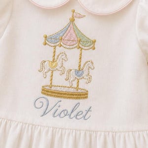 May include: A white short-sleeved dress with a Peter Pan collar featuring pink trim. The chest displays an embroidered carousel with horses in pastel colours and the name Violet stitched in blue thread below the carousel design.
