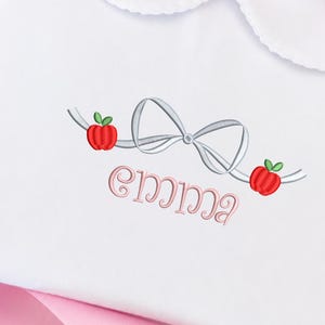 May include: A white shirt with a rounded Peter Pan collar featuring an embroidered design of a silver ribbon bow flanked by two red apples with green leaves. Below the ribbon, the name Emma is embroidered in a light pink cursive script font.