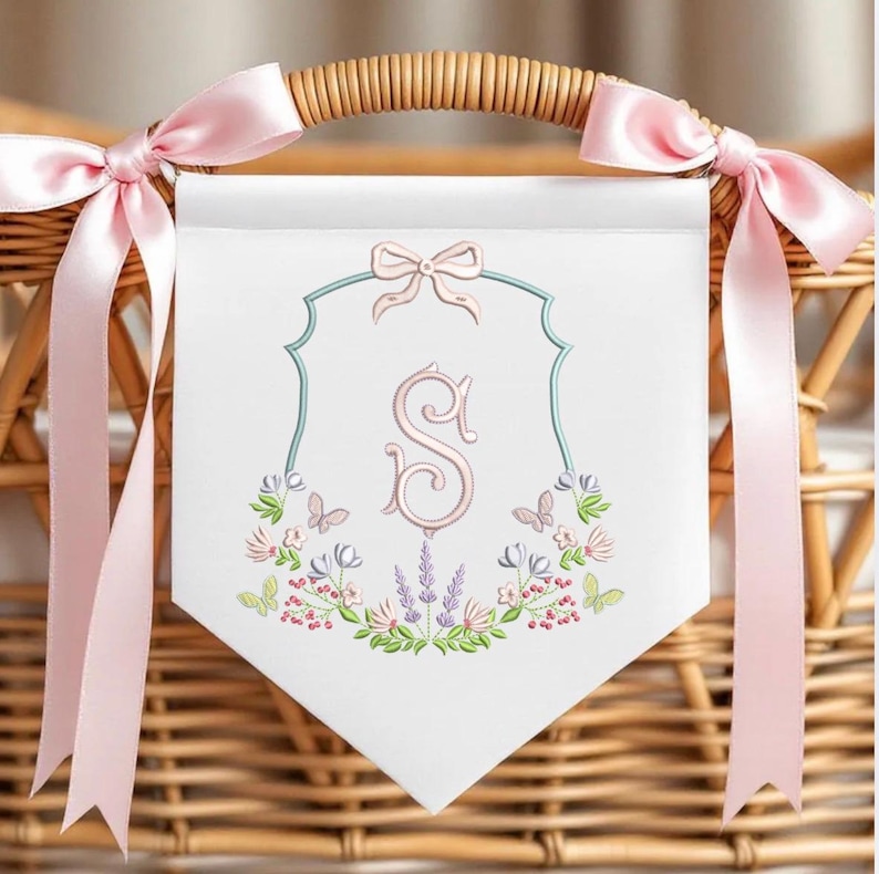 May include: White banner with a pink bow and the letter "S" embroidered in pink. The banner is decorated with butterflies and floral embroidery, and is tied with pink ribbons to a wicker basket.