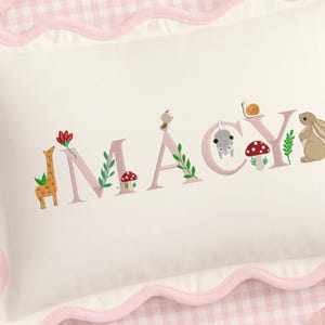 Op de afbeelding: Een wit kussen met een roze geschulpte rand, geborduurd met de naam "MACY" in roze letters. Elke letter is versierd met bosdieren en bloemmotieven, waaronder een giraf, paddenstoel en konijn.