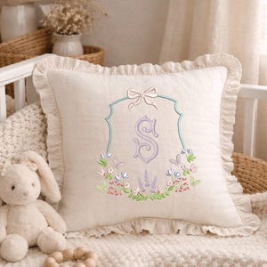 May include: A cream-colored decorative pillow with a ruffled edge. The pillow features an embroidered design of the letter "S" in a lavender color, framed by a light blue border and a bow at the top. Floral and butterfly embroidery surrounds the letter.