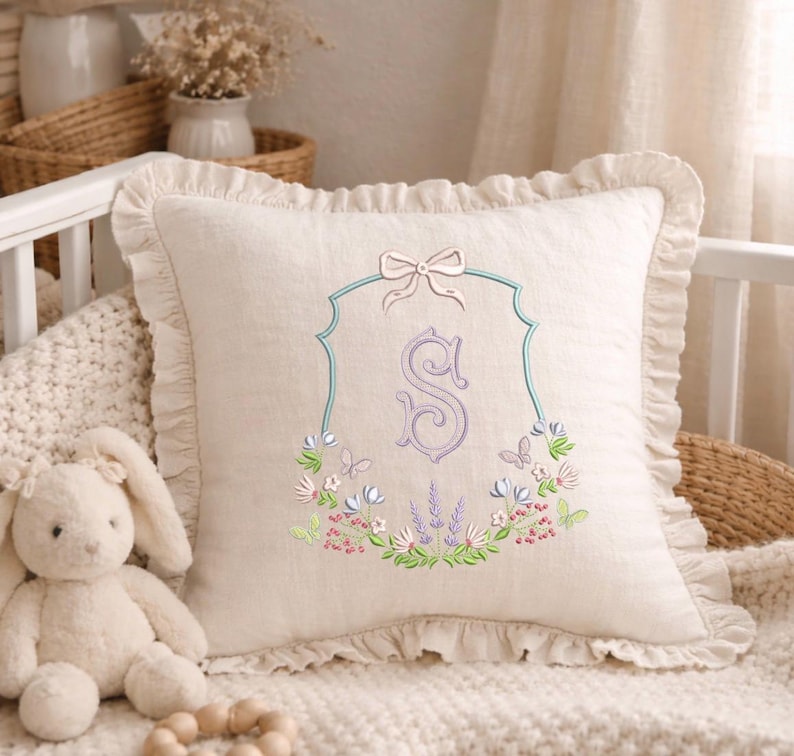 May include: A cream-colored decorative pillow with a ruffled edge. The pillow features an embroidered design of the letter "S" in a lavender color, framed by a light blue border and a bow at the top. Floral and butterfly embroidery surrounds the letter.
