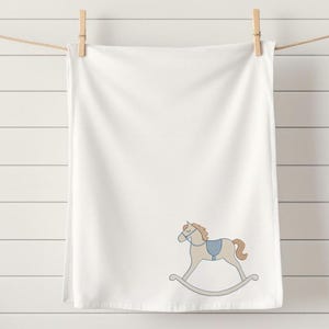 Puede incluir: Toalla blanca con un bordado de caballo balanc&iacute;n en azul claro y beige. La toalla est&aacute; colgada de un tendedero con una pinza de madera. El fondo es una pared de listones blancos.
