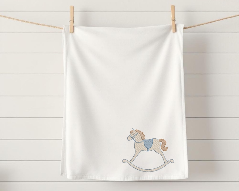 Puede incluir: Toalla blanca con un bordado de caballo balanc&iacute;n en azul claro y beige. La toalla est&aacute; colgada de un tendedero con una pinza de madera. El fondo es una pared de listones blancos.