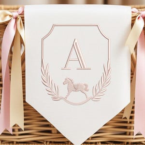 Può includere: Un banner bianco con un ricamo color oro rosa raffigurante la lettera "A", un cavallo a dondolo e foglie di alloro. Lo stendardo è decorato con nastri rosa e crema, il tutto su un cesto di vimini.