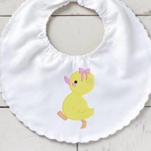 Puede incluir: Babero blanco para bebé con borde festoneado y pato bordado amarillo con lazo rosa. El pato tiene pico y patas rosas. El babero tiene cierre de lazo en el cuello.
