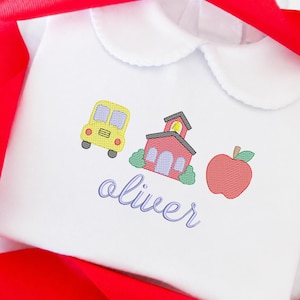 May include: A white collared shirt featuring an embroidered design of a yellow school bus, a red schoolhouse, and a red apple. Below the icons, the name oliver is embroidered in light blue cursive script. The shirt is framed by red fabric.