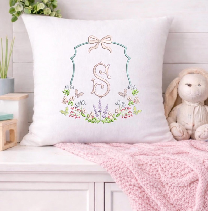 May include: White square pillow with an embroidered floral design and the letter "S". The design includes a light blue frame, a bow, butterflies, and various flowers in shades of pink, green, and purple. A pink blanket is draped below.