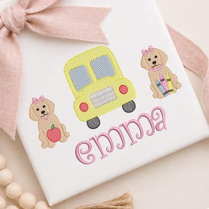 May include: White fabric featuring embroidered school bus, puppies, and the name "Emma" in pink. The yellow school bus has black wheels and red lights. The puppies have pink bows and are holding an apple and crayons.