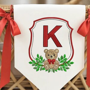 May include: White banner with a red embroidered letter "K" inside a shield design. A teddy bear with a red bow tie and green leaves are embroidered below the letter. Red ribbons are tied on each side.