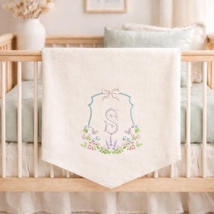 May include: Cream-colored baby blanket with an embroidered floral design and the letter "S". The embroidery features a light blue frame, a pink bow, and a wreath of flowers in pink, purple, and green. The blanket is draped over a wooden crib.