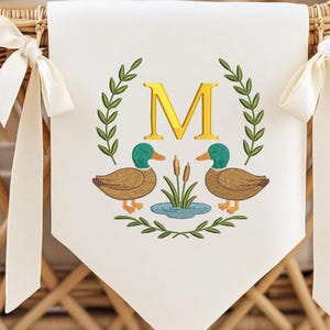 Puede incluir: Una pancarta blanca con un diseño bordado de dos patos, un estanque y la letra "M" en oro. La pancarta está atada con cintas de color crema.