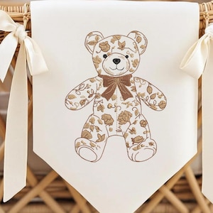 May include: A cream banner featuring a brown embroidered teddy bear with floral patterns and a bow tie. The banner is attached to a wicker basket with cream ribbons tied on each side. The design is suitable for a nursery.