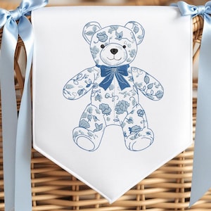 Op de afbeelding: Een witte banner met een blauw bloemen teddybeer ontwerp. De beer heeft een blauwe strik en wordt omringd door blauwe linten. De banner hangt voor een rieten mand.