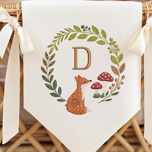 Puede incluir: Bandera crema con un diseño bordado de bosque. Presenta un zorro, setas y la letra "D" dentro de una corona de hojas. Cintas color crema atan la bandera a una cesta de mimbre.