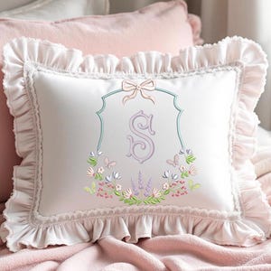 May include: White decorative pillow with a ruffled edge and a floral embroidered design. The pillow features a light pink bow and a monogrammed letter "S" in a decorative frame. The pillow is on a pink blanket.