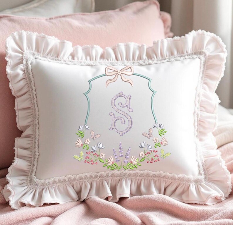 May include: White decorative pillow with a ruffled edge and a floral embroidered design. The pillow features a light pink bow and a monogrammed letter "S" in a decorative frame. The pillow is on a pink blanket.