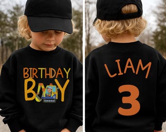 May include: Black sweatshirt with "BIRTHDAY BOY" text and a construction vehicle graphic. The back of the sweatshirt has the name "LIAM" and the number "3" in orange. The child is wearing a black baseball cap.