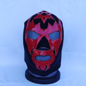 Puede incluir: Una máscara de lucha libre negra y roja con un diseño de pentagrama en la parte superior. La máscara tiene una cara roja con ojos negros y una barba negra.